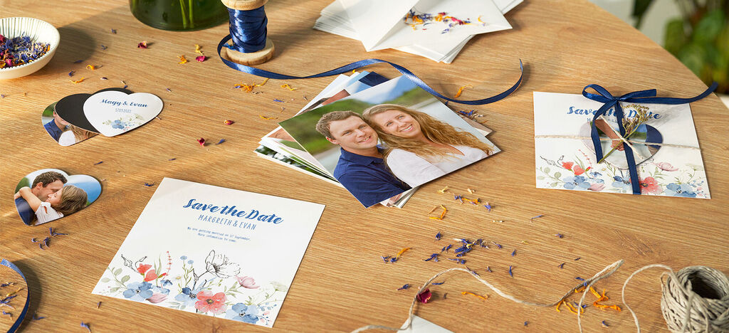 A table filled with wedding decorations. Various greeting cards, photos, and scatter decor are lovingly arranged. The cards display 'Save the Date' in a blooming design.
