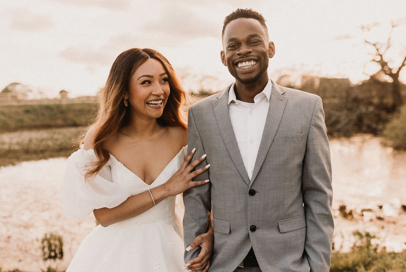 A bride and groom stand near a body of water and smile. The bride wears an elegant white gown and the groom a gray suit.