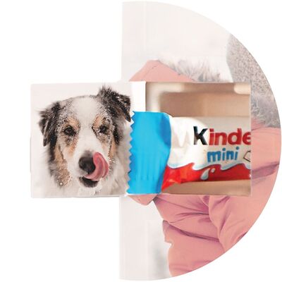 A dog with wet fur licks its nose while standing next to a colorful Kinder chocolate packaging
