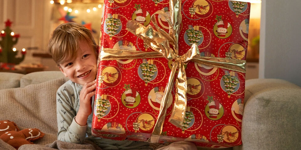 A child holds a large, lovingly wrapped gift with festive paper in their hands. The room is beautifully decorated for Christmas.