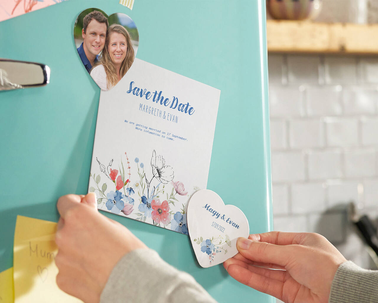 A hand attaches a "Save the Date" card with floral motifs to a fridge.