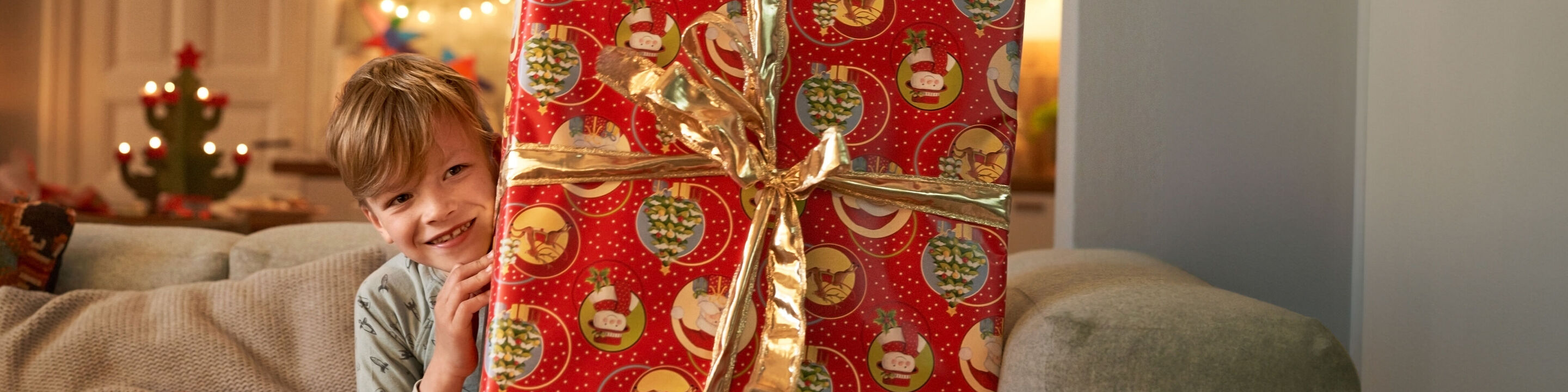 A festively wrapped gift in red paper with a golden ribbon. Background features glowing Christmas lights.