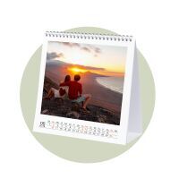 A couple sits on a cliff watching the sunset over the sea, accompanied by a CEWE photo calendar featuring this image.