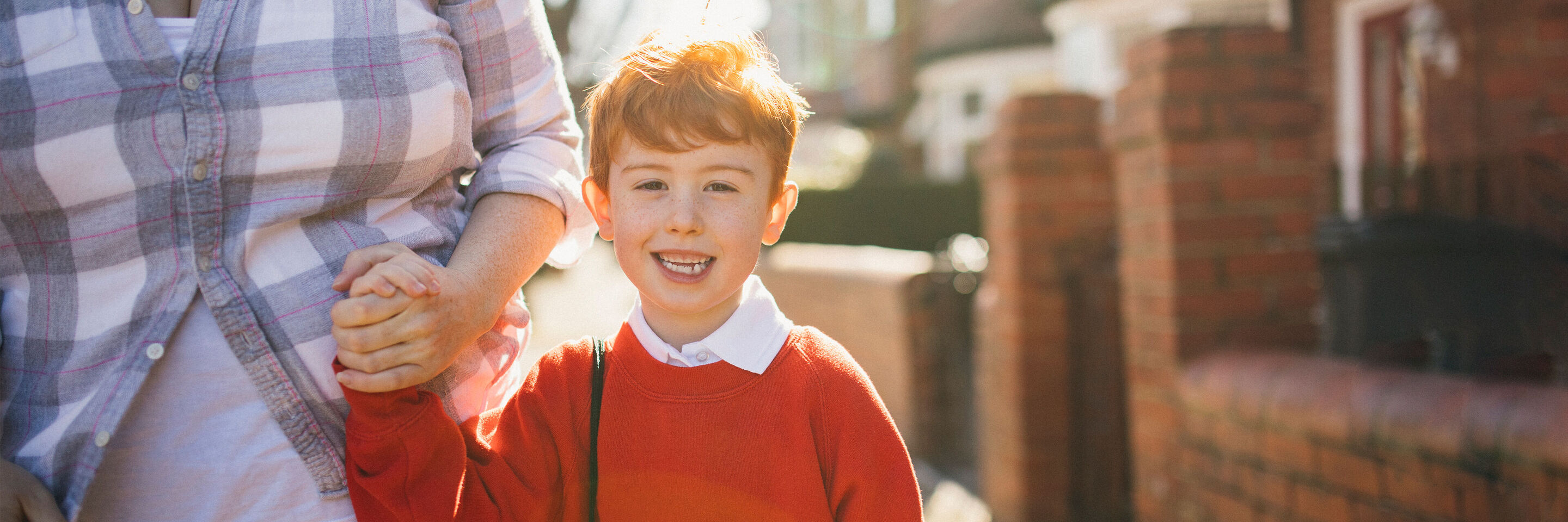 A child in a red sweater holds an adult's hand and smiles. They walk together along a sunny street.