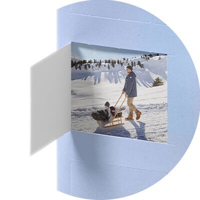A man pulls a friend on a sled through the snow. Snowy mountains and a blue sky are in the background. It's a cheerful winter day.