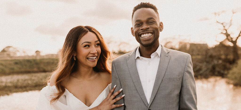 A couple in elegant clothing stands by the water and smiles.