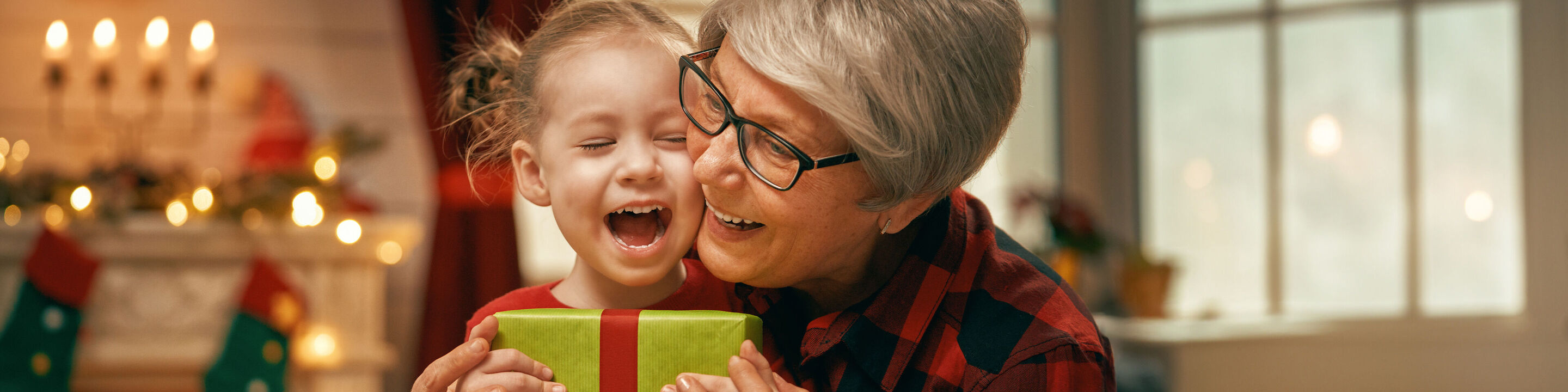 A child and an adult hold a colorful gift package together, surrounded by festive Christmas decorations.
