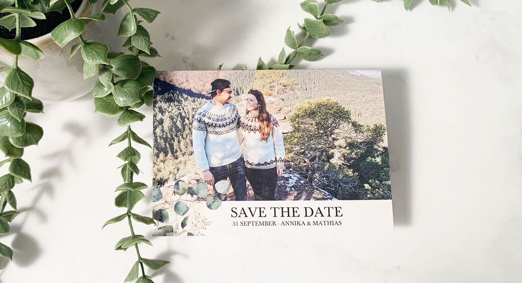 A couple in sweaters smiles in front of a mountain landscape, surrounded by plants. It's a wedding invitation.